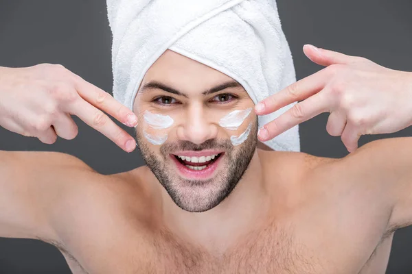Усміхнений бородатий чоловік в рушнику наносить крем для обличчя, ізольований на сірому, концепція догляду за шкірою — стокове фото