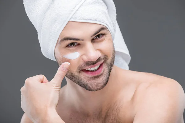 Barbudo alegre en toalla aplicando crema facial, aislado en gris, concepto de cuidado de la piel - foto de stock