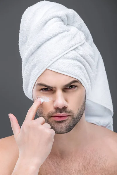 Homem em toalha aplicando creme cosmético no rosto, isolado em cinza, conceito de cuidados com a pele — Fotografia de Stock