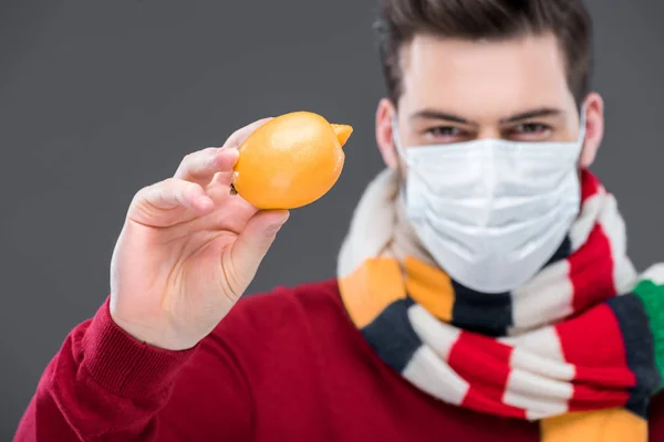 Больной человек в теплом шарфе и медицинской маске держит лимон, изолированный на сером — стоковое фото