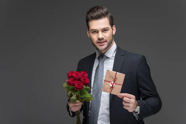 Schöner Mann im Anzug mit roten Rosen und Umschlag zum Valentinstag, isoliert auf grau — Stockfoto