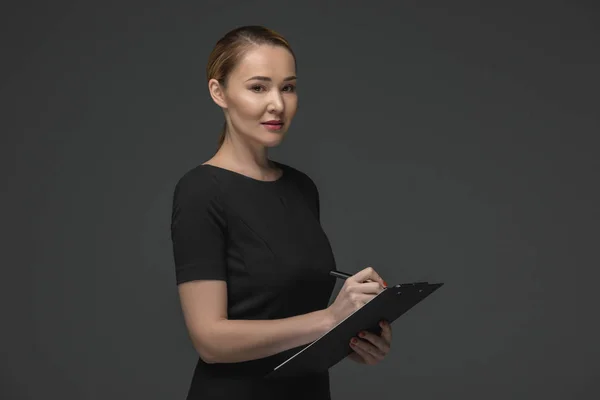 Smiling kazakh businesswoman writing on clipboard and looking at camera isolated on grey — Stock Photo