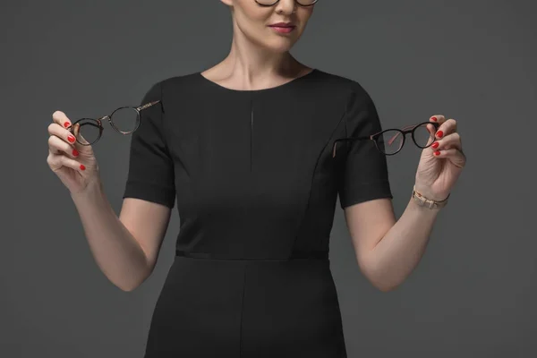 Tiro cortado de mulher em vestido preto escolhendo óculos isolados em cinza — Fotografia de Stock