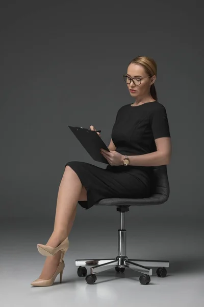 Hermosa mujer de negocios kazakh serio escribir en portapapeles en gris - foto de stock