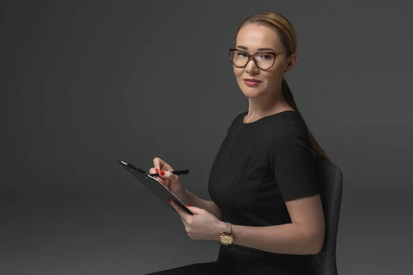 Hermosa mujer de negocios kazakh escribir en el portapapeles y mirando la cámara aislada en gris - foto de stock