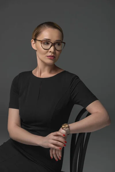 Belle femme kazakh dans les lunettes assis sur la chaise et regardant loin isolé sur gris — Photo de stock