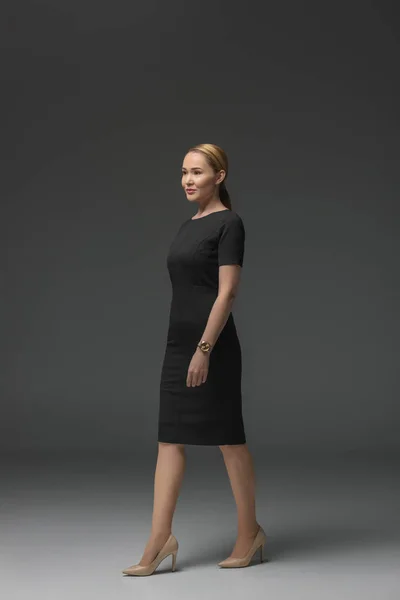 Visão comprimento total da bela mulher kazakh em vestido preto andando e olhando para longe em cinza — Fotografia de Stock