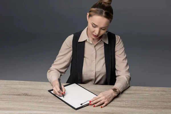 Attraktive kasachische Geschäftsfrau schreibt auf Klemmbrett am Tisch isoliert auf grau — Stockfoto