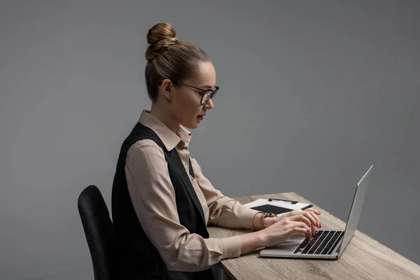 Vista lateral da empresária kazakh em óculos usando laptop na mesa isolada em cinza — Fotografia de Stock