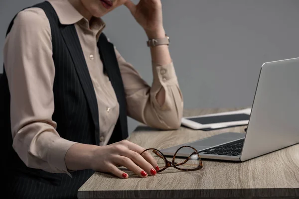 Recortado disparo de mujer de negocios sosteniendo gafas mientras se utiliza el ordenador portátil en la mesa en gris - foto de stock