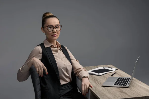 Приваблива казахська бізнес-леді в окулярах використовує ноутбук і дивиться на камеру ізольовано на сірому — стокове фото