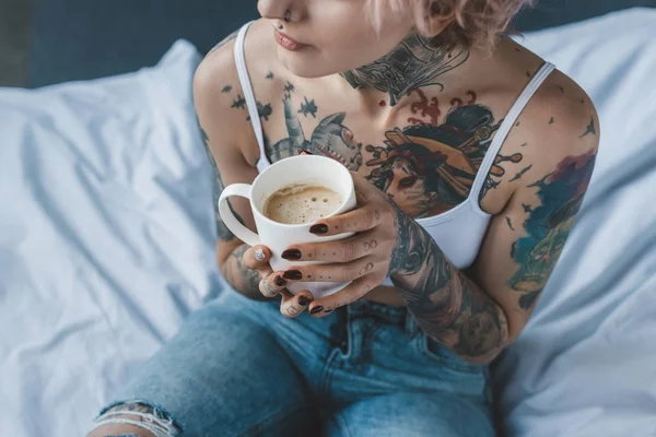 Vista recortada de chica tatuada con pelo rosa bebiendo café en la cama por la mañana - foto de stock