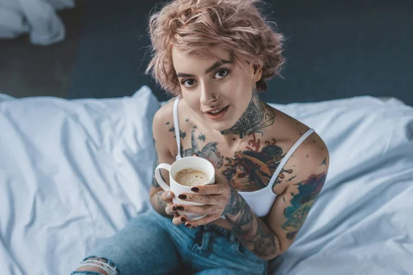 Young tattooed girl with pink hair drinking coffee in bed at morning — Stock Photo