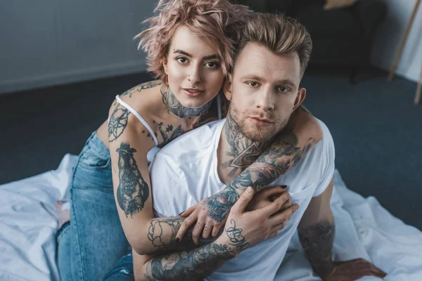 Tattooed girlfriend looking at camera and hugging her boyfriend on bed in bedroom — Stock Photo