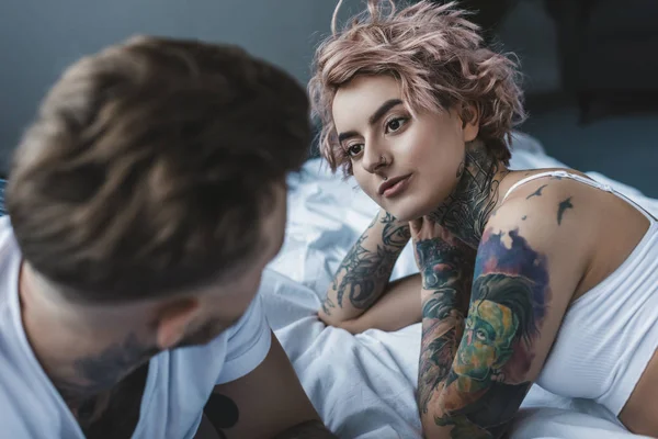 Jeune couple tatoué regardant l'autre et couché sur le lit dans la chambre — Photo de stock