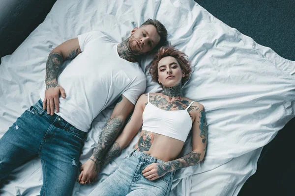 Vista superior de la joven pareja tatuada cogida de la mano y acostada en la cama - foto de stock