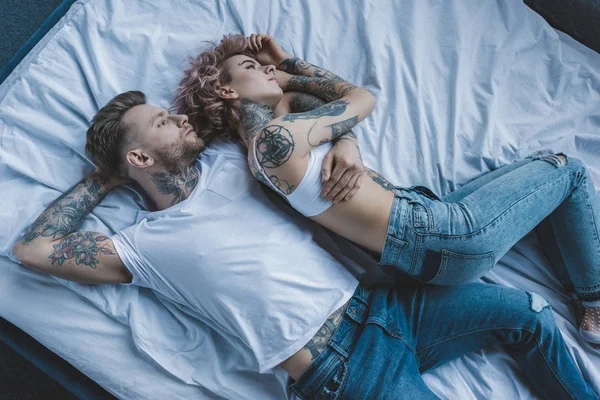 Vista superior do casal tatuado abraçando e deitado na cama no quarto — Fotografia de Stock