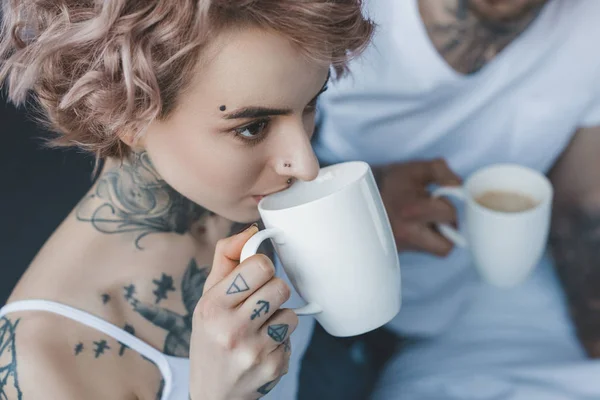 Joven chica tatuada bebiendo café por la mañana, novio sentado detrás - foto de stock