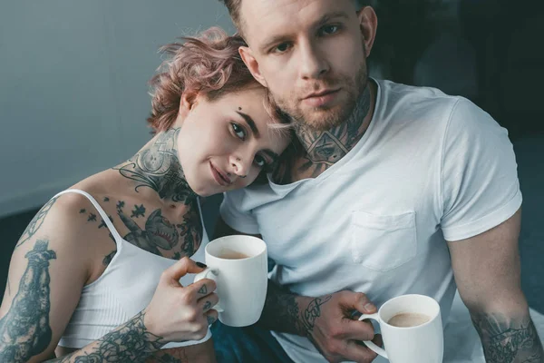 Молодая татуированная пара смотрит в камеру и пьет кофе в постели по утрам — Stock Photo