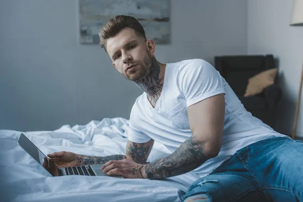 Hombre tatuado guapo mirando a la cámara mientras usa el ordenador portátil en la cama - foto de stock