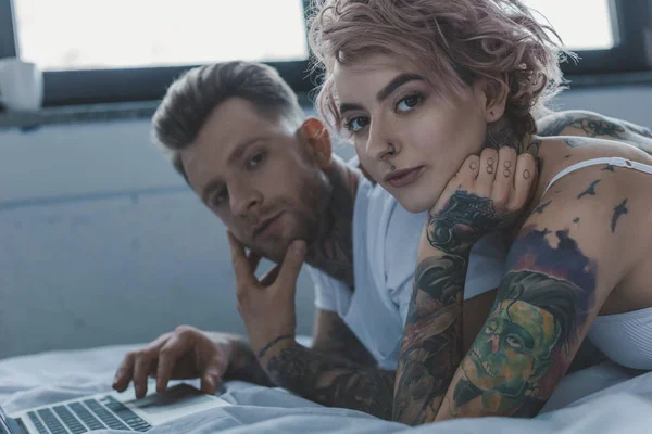 Joven pareja tatuada usando portátil en la cama - foto de stock