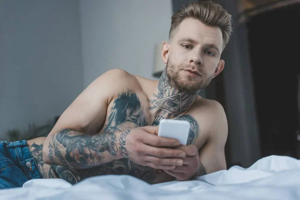 Bel homme tatoué torse nu utilisant un smartphone sur le lit — Photo de stock