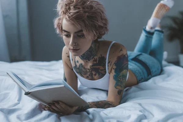 Молодая девушка с татуировками с розовыми волосами читает книгу, лежа на кровати — стоковое фото