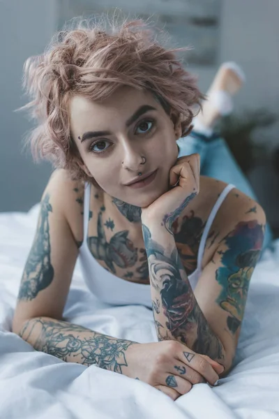 Chica tatuada con el pelo rosa acostado en la cama y mirando a la cámara - foto de stock