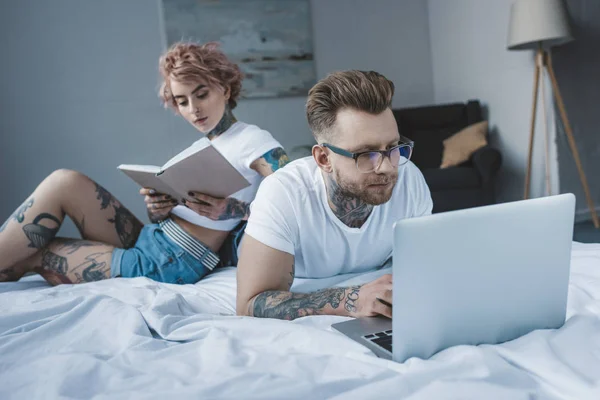 Novia tatuada leyendo libro mientras novio usando portátil en la cama - foto de stock