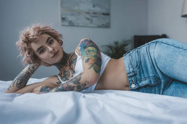 Belle fille encrée avec des cheveux roses couché sur le lit et regardant la caméra — Photo de stock