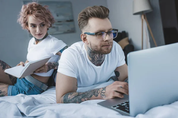 Livre de lecture de couple tatoué et en utilisant un ordinateur portable au lit — Photo de stock
