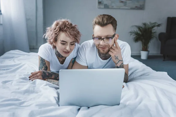 Татуированная улыбающаяся пара, использующая ноутбук в постели — стоковое фото