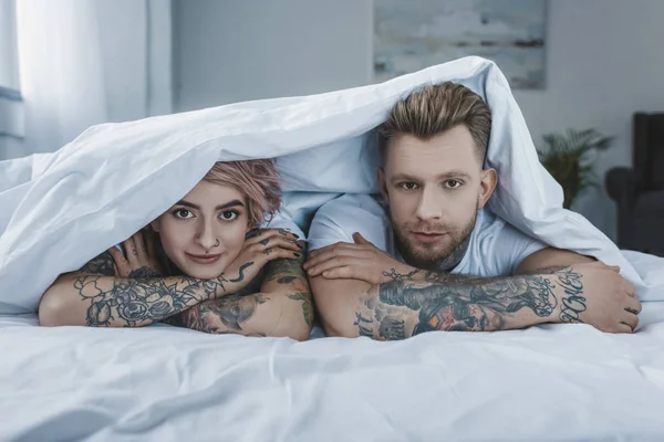 Giovane coppia tatuata sdraiata a letto sotto una coperta bianca — Foto stock