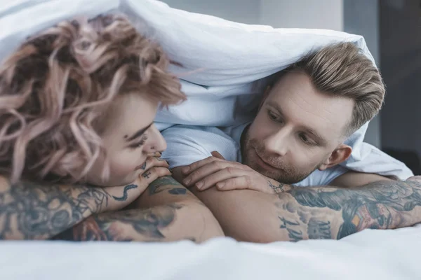 Young tattooed couple looking at each other under white blanket in bedroom — Stock Photo