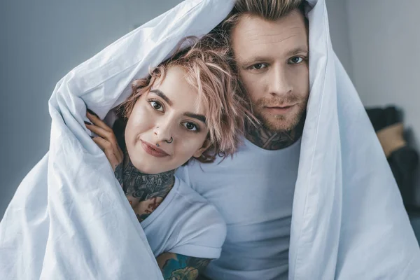 Joven pareja tatuada mirando la cámara debajo de una manta blanca en el dormitorio - foto de stock