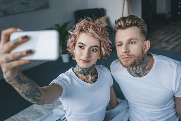 Beautiful tattooed couple taking selfie on smartphone in bedroom — Stock Photo