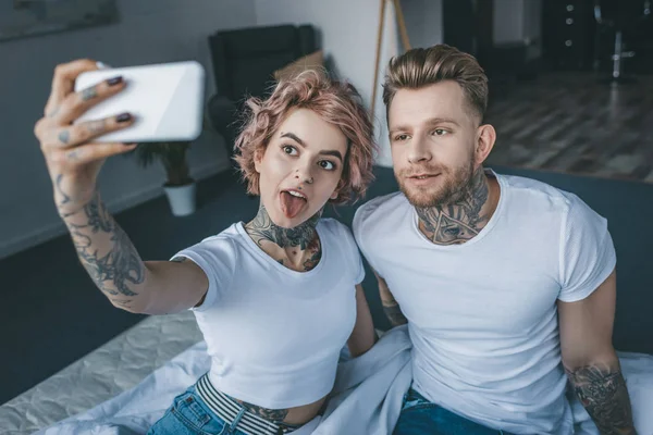 Hermosa chica entintada pegando la lengua y tomando selfie con novio en el dormitorio - foto de stock