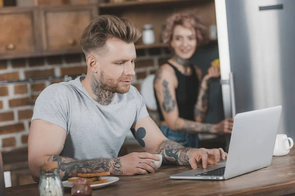 Tatuado homem usando laptop durante pequeno-almoço, namorada atrás na cozinha — Fotografia de Stock