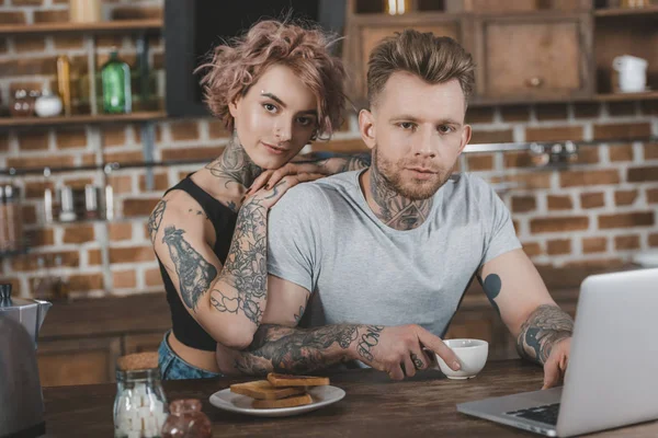 Татуированная пара с помощью ноутбука и завтраком на кухне — стоковое фото