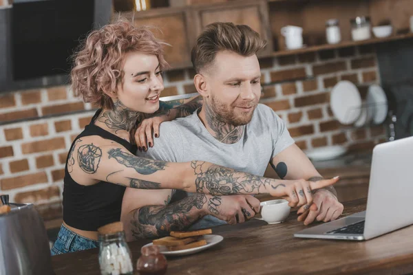 Tattooed couple pointing at laptop during breakfast in kitchen — Stock Photo