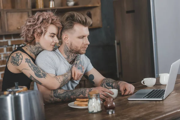 Татуированная пара обнимается и использует ноутбук во время завтрака на кухне — стоковое фото