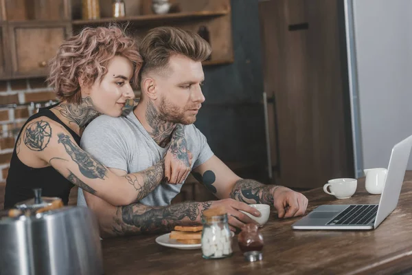 Pareja tatuada abrazando y utilizando el ordenador portátil durante el desayuno en la cocina - foto de stock