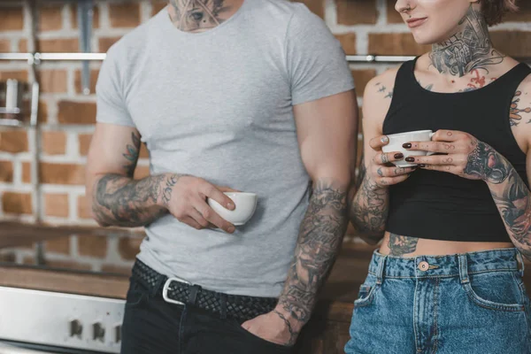 Cropped view of tattooed couple drinking coffee in kitchen at morning — Stock Photo