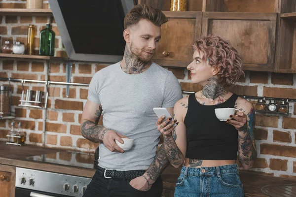 Young tattooed couple drinking coffee and using smartphone in kitchen at morning — Stock Photo