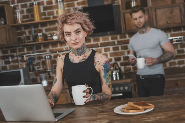 Fille tatouée à l'aide d'un ordinateur portable pendant le petit déjeuner, petit ami derrière la cuisine — Photo de stock