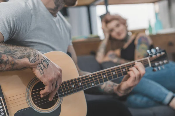 Tätowierter Freund spielt zu Hause für seine Freundin auf der Akustikgitarre — Stockfoto