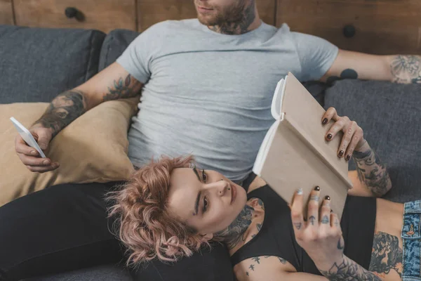 Novia leyendo libro mientras novio usando smartphone en casa - foto de stock