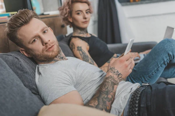Татуированная пара, использующая смартфон и ноутбук дома — стоковое фото