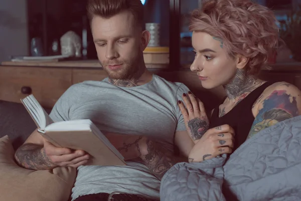 Casal tatuado descansando e lendo livro em casa — Fotografia de Stock