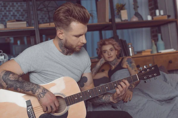 Novio tatuado tocando en la guitarra acústica para su novia en casa - foto de stock